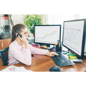 A SupremeX employee sits at a desk in a professional office, analyzing financial or business data displayed on two monitors. She is engaged in a phone call while typing on a keyboard, indicating a role in finance, analytics, or business management.