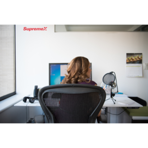 A SupremeX employee sits at a modern office desk, facing a computer screen. The workspace includes a microphone, headphones, and neatly organized documents, suggesting a role in communication, customer service, or remote work.