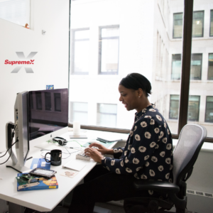 A professional woman with short black hair, wearing a dark floral blouse, is sitting at a modern office desk typing on a keyboard. She is smiling while working on a task. The desk is organized with a computer monitor, a notebook, a coffee mug, and some books. A large window behind her offers a view of an urban office building. The "Supremex" logo is visible on the wall.