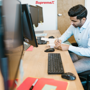 A professional man with short dark hair, wearing a light blue dress shirt, is seated at a modern office desk, writing on a notepad. Multiple computer monitors, keyboards, and office supplies such as folders and documents are spread across the wooden desk. The "Supremex" logo is visible on the wall, and a closed wooden door is in the background. The office environment appears organized and professional.